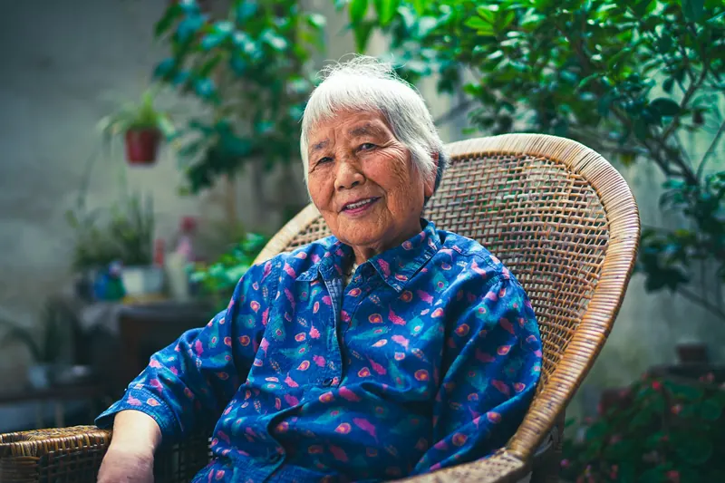 Smiling older Asian woman sitting in a wicker chair on a leafy patio